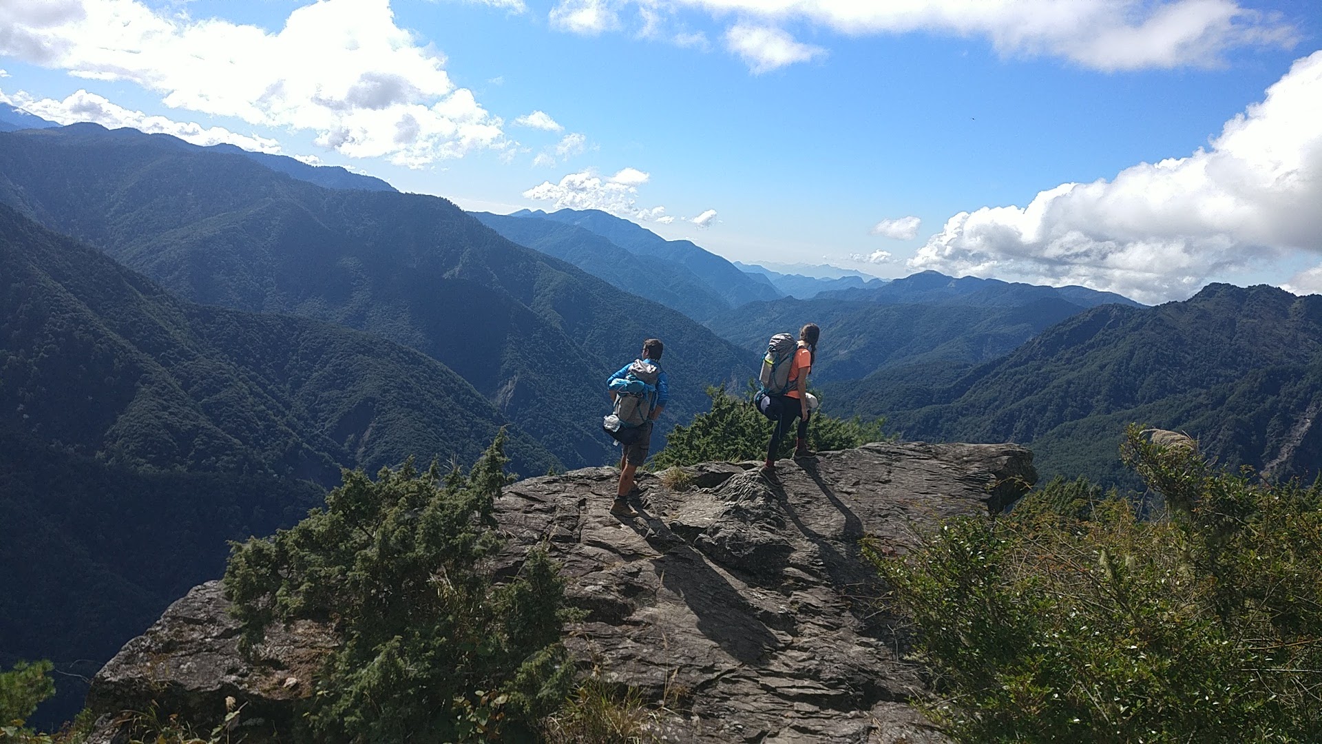 Two people on a rock, with hicking backpacks and stunning mountain scenery behind